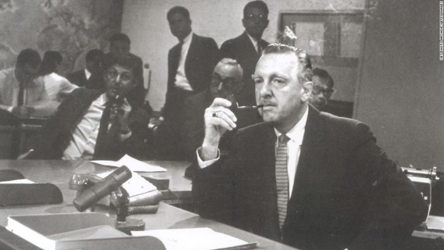 Walter Cronkite sits and smokes a pipe at the CBS news desk with Don Hewitt in the background.